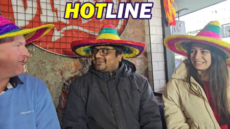 Kali, an Indian chef, sits between Sarah Jane Seddon and Sean Rush during a spontaneous interview on Hotline Folks. All three are wearing colorful sombreros and smiling, creating a lighthearted atmosphere. The setting reflects the casual and fun tone of the conversation, with a red sign in the background adding to the vibrant scene.